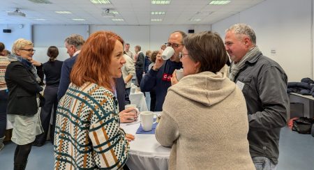 Mehrere Personen stehen in einem hellen Raum an Stehtischen und unterhalten sich, einige halten Tassen in der Hand.