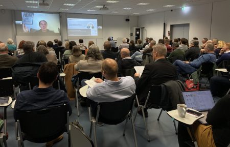 Ein Vortragssaal mit mehreren Personen, die auf einen Bildschirm an der Vorderseite des Raums schauen. Die meisten sitzen auf schwarzen Stühlen mit kleinen Tischen, einige haben Notizen oder Laptops vor sich. An der Wand ist eine Präsentation mit Text und einem Bild zu sehen. Die Decke ist mit quadratischen Leuchtpaneelen ausgestattet, und die Wände sind weiß.