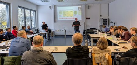 Referent steht vor Projektionsleinwand und spricht zu einer Gruppe von Erwachsenen, die an Tischen sitzen und zuhören.