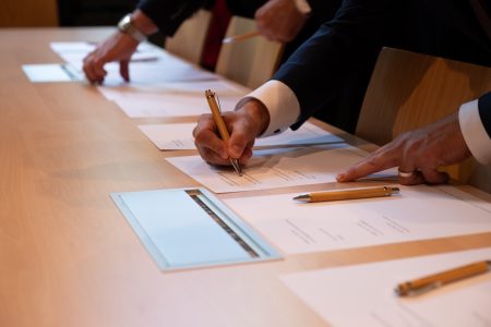 Auf einem hölzernen Schreibtisch liegen die Urkunden. Eine Hand, in weißem Hemd und dunkelblauem Sakko, unterschreibt mit einem hölzernen Kulli die Urkunde.