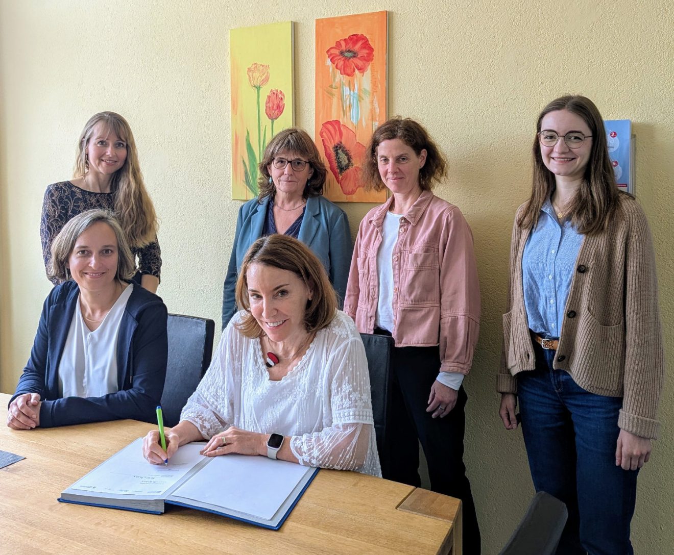 Die Rektorin unterzeichnet die Vereinbarung. Um sie herum sitzen und stehen die fünf weiteren Teilnehmerinnen. Alle lächeln erfreut.