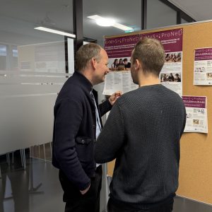 Zwei Projekt- Mitarbeitende stehen vor einer Posterwand zu dem KuMuS-Projekt und unterhalten sich angeregt