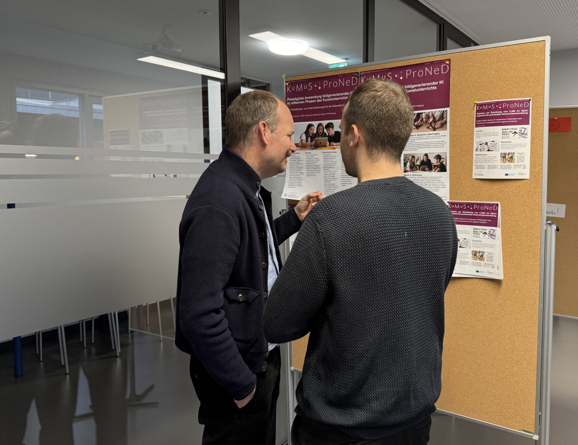 Zwei Projekt- Mitarbeitende stehen vor einer Posterwand zu dem KuMuS-Projekt und unterhalten sich angeregt