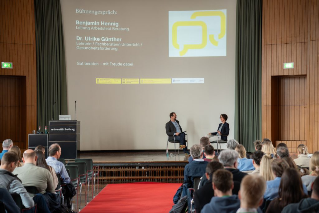 Blickt auf die Bühne der gut gefüllten Aula, wo zwei Teilnehmer*innen im Podiumsgespräch sind