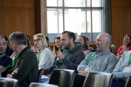 Blick ins Publikum während des Austauschs mit Andreas Kronenberg. Ein junger Mann mit grauen Hoodie und grünen Tagungsband um gehängt, hält ein Mikrofon in der Hand und stellt gerade eine Frage.