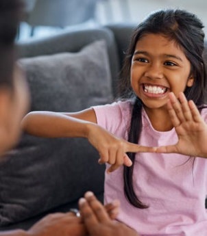 Kind mit Zöpfen zeigt mit den Fingern in deutscher Gebärdensprache eine Zahl vor einer anderen Person auf einem Sofa