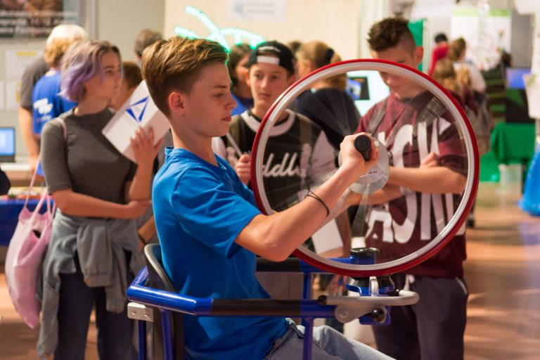 Eine Gruppe Schülerinnen und Schüler schauen interessiert einem weiteren Schüler zu, wie er für ein physikalisches Experiment an einem Fahrradrad dreht.