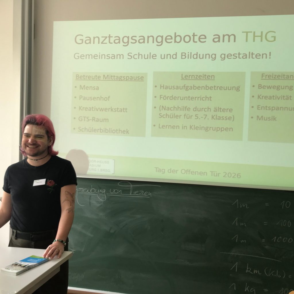Ein junger Mann steht an einem Pult, im Hintergrund sieht man eine Präsentation zum Thema Ganztagsangebote am THG