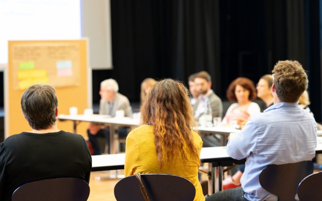 Man sieht Menschen, die an Tischen sitzen und folgen einem Gespräch. Vorne sind Stellwände mit Notizzetteln zu sehen.