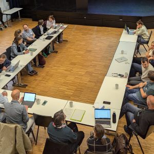 Blick von oben in die Aula, wo die 17 Teilnehmenden des Treffens zusammen sitzen und sich austauschen