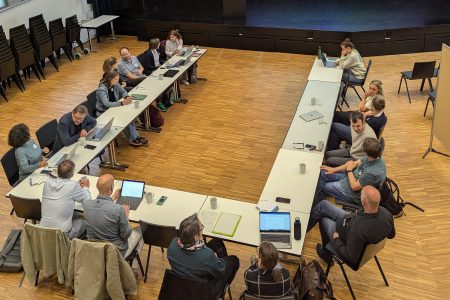 Blick von oben in die Aula, wo die 17 Teilnehmenden des Treffens zusammen sitzen und sich austauschen