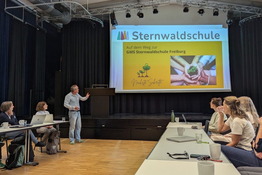 Marc Joos, Schulleiter der Sternwaldschule, steht vor den Teilnehmenden in der Aula und berichtet mit einer Präsentation von seiner Schule