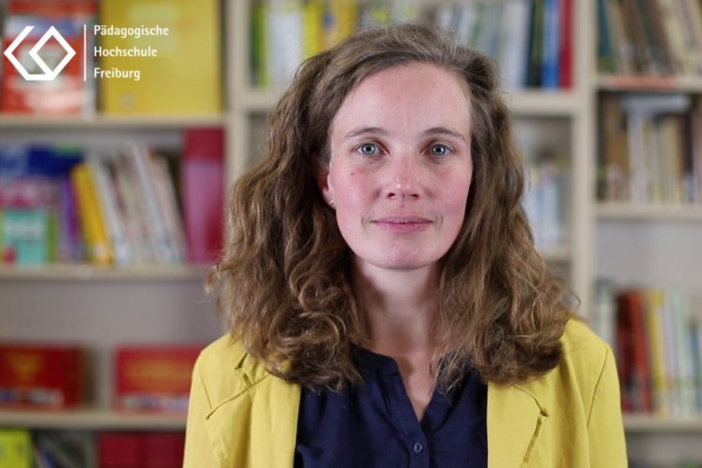 Eine Wissenschaftlerin mit einem gelben Blazer und einer blauen Bluse steht vor einer Bücherwand und blickt direkt in die Kamera. Es handelt sich um einen Screenshot eines F3 Nugget-Films.