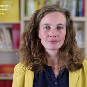 Eine Wissenschaftlerin mit einem gelben Blazer und einer blauen Bluse steht vor einer Bücherwand und blickt direkt in die Kamera. Es handelt sich um einen Screenshot eines F3 Nugget-Films.