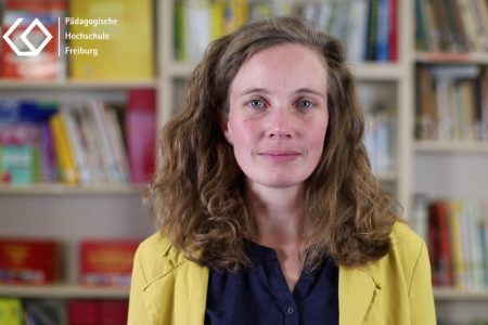 Eine Wissenschaftlerin mit einem gelben Blazer und einer blauen Bluse steht vor einer Bücherwand und blickt direkt in die Kamera. Es handelt sich um einen Screenshot eines F3 Nugget-Films.