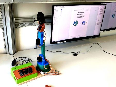 Roboterarm mit mehreren Gelenken und Kabeln auf einem Tisch vor einem Monitor mit geöffneter Softwareoberfläche