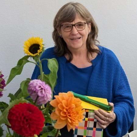 Barbara Skorupsinki trägt ein blaues Oberteil und hält lächelnd ein bunt verpacktes Geschenk in den Händen. Neben ihr steht ein bunter Blumenstrauß.