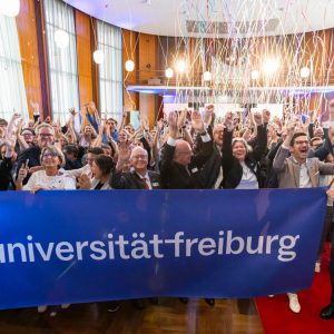Zahlreiche Mitglieder der Universität Freiburg halten jubelnd einen Banner mit dem Logo der Universität hoch, während im Hintergrund der Aula bunte Luftschlangen durch die Luft fliegen