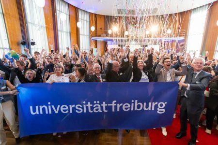Zahlreiche Mitglieder der Universität Freiburg halten jubelnd einen Banner mit dem Logo der Universität hoch, während im Hintergrund der Aula bunte Luftschlangen durch die Luft fliegen
