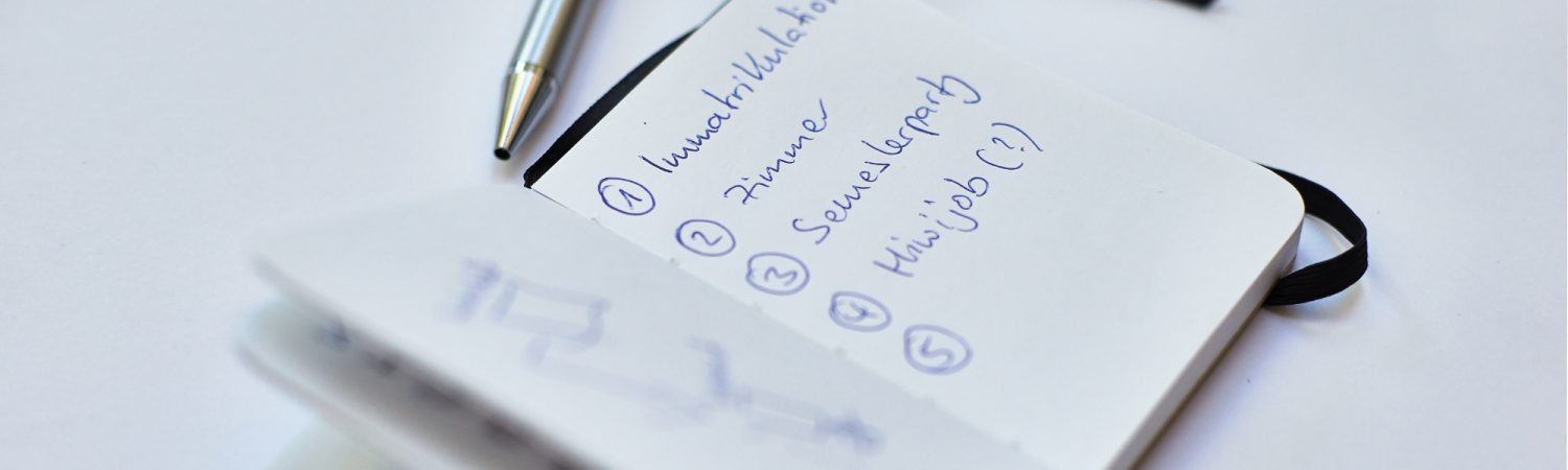 In einem aufgeschlagenen Notizheft sind die Punkte 1: Immatrikulation, 2: Zimmer, 3: Semesterparty und 4: Minijob (?) notiert. Neben dem Notizheft liegt ein Kulli