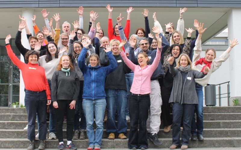 Eine Gruppe von mehr als 25 Personen steht fröhlich auf den Treppen vor dem PH Gebäude und viele strecken ihre Hände in die Luft